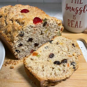 May include: A loaf of fruitcake with a slice cut off, revealing raisins and candied fruit. The cake is topped with red cherries. A white mug with the text "THE SNUGGLE IS REAL" is in the background.