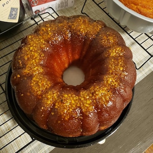 May include: A glazed bundt cake with a brown sugar glaze and chopped nuts on top. The cake is on a black cooling rack.