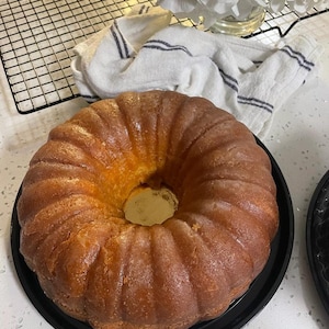 May include: A golden brown bundt cake with a glazed finish, sitting on a black round cake plate. The cake is on a white countertop.