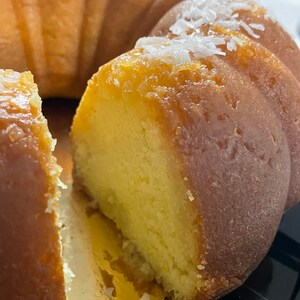 May include: A close-up of a golden yellow bundt cake with a slice removed, revealing the moist, light yellow interior. The cake is topped with white coconut flakes.