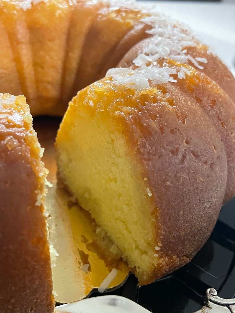 May include: A close-up of a golden yellow bundt cake with a slice removed, revealing the moist, light yellow interior. The cake is topped with white coconut flakes.