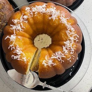 May include: A yellow bundt cake with white coconut flakes on top, sitting on a black glass cake stand. A silver cake server with the text "I'm just here for dessert" is on the stand.