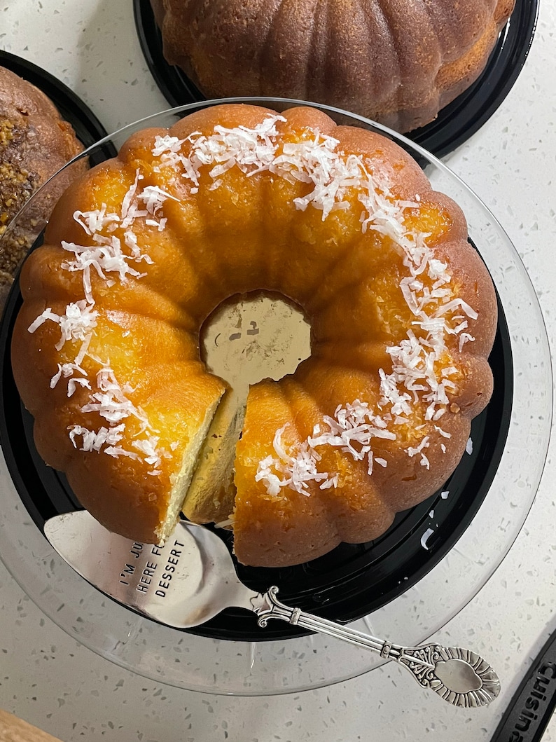 May include: A yellow bundt cake with white coconut flakes on top, sitting on a black glass cake stand. A silver cake server with the text "I'm just here for dessert" is on the stand.