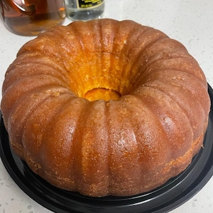 May include: A golden-brown bundt cake with a glossy glaze sits on a black plate. The cake has a ring shape with a textured, ridged surface. The cake is a dessert item, possibly for a special occasion.