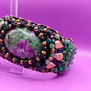 Pode incluir: Uma pulseira de contas verdes, rosa e pretas com uma grande pedra preciosa verde e rosa no centro. A pulseira é decorada com contas e detalhes dourados.