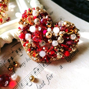 May include: A heart-shaped brooch made of red, gold, and white beads. The brooch is decorated with pearls, rhinestones, and gold accents. The brooch is lying on a sheet of music.