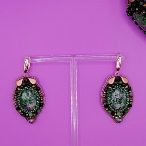 May include: A pair of dangle earrings with a leaf-like design. The earrings feature a central oval stone with green and white marbling, surrounded by a beaded border in shades of green, black, and purple. Gold-colored metal accents are visible.