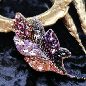 May include: A delicate leaf-shaped brooch with intricate beadwork. The brooch features a gradient of colors, starting with deep purple and transitioning to pink and gold. The brooch is adorned with sparkling crystals and beads.