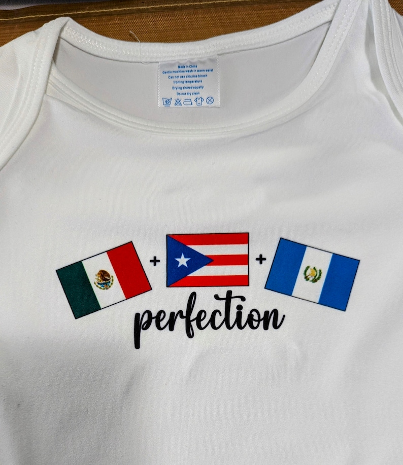 May include: White baby onesie with three flags printed on it. The flags are the Mexican flag, the Puerto Rican flag, and the Guatemalan flag. The text "perfection" is printed below the flags.
