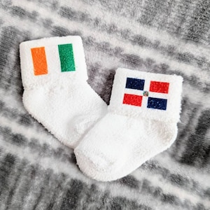 May include: Two white fuzzy socks with flags printed on them. One sock has the Irish flag, which is green, white, and orange. The other sock has the Dominican Republic flag, which is red, white, and blue.