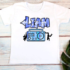 May include: White t-shirt with a blue and white graphic of a boombox and the name "LIAM" in a graffiti style font.