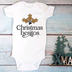 May include: White baby onesie with a gold holly leaf design and the text "Christmas besitos".
