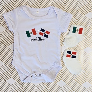 May include: White baby onesie with a graphic of two flags, a Mexican flag and a Dominican Republic flag, with the text " + perfection". The onesie is paired with a pair of white baby socks, one with a Mexican flag and the other with a Dominican Republic flag.