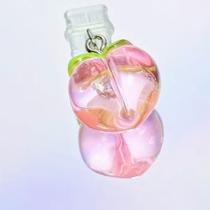 May include: A clear glass bottle stopper with a peach-shaped charm. The charm is a translucent pink with green leaves and a silver-colored metal loop. The stopper is designed to fit into a bottle opening.