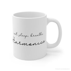 May include: White ceramic mug with the text "eat, sleep, breathe Harmonica" in black script. The mug has a curved handle and a glossy finish. The words are arranged in a vertical format.