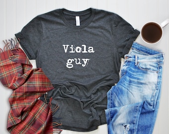 May include: A dark gray t-shirt with the text "viola guy" printed in white on the front. The shirt is laid flat on a white surface with a plaid scarf and blue jeans.