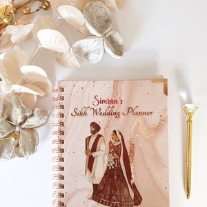 May include: A pink and gold marble-patterned notebook with a gold spiral binding. The cover features an illustration of a Sikh couple in traditional wedding attire. The text on the cover reads "Simran's Sikh Wedding Planner" and "By Kismat Weddings".