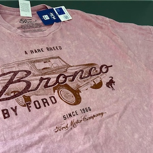 May include: Pink t-shirt featuring a distressed graphic of a Ford Bronco SUV. The design includes the text "A Rare Breed," "Bronco by Ford," and "Since 1966 Ford Motor Company." The shirt has a vintage, faded look.