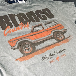 May include: Gray t-shirt featuring an orange vintage Bronco SUV graphic. The text "Bronco Country" is above the vehicle, with "Ford Motor Company Since 1966" printed below. The shirt has a vintage, worn look.