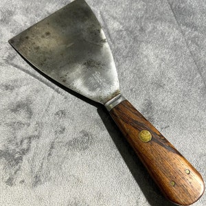 May include: A vintage putty knife with a wooden handle and brass accents. The blade is made of metal and has a slight curve. The handle is dark brown with a wood grain pattern. The blade is stamped with the text "The Stanley Works" and "P.T.A."