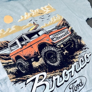 May include: Light blue t-shirt featuring an orange Bronco SUV with a surfboard on top, set against a beach scene with a sun and clouds. The word "Bronco" is prominently displayed, along with the Ford logo.