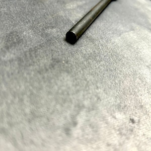 May include: A close-up of a long, thin, silver metal rod with a rounded end. The rod is lying on a gray surface.