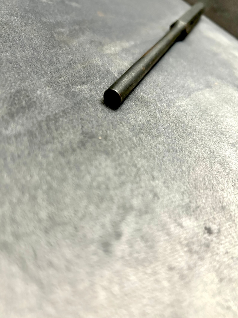 May include: A close-up of a long, thin, silver metal rod with a rounded end. The rod is lying on a gray surface.