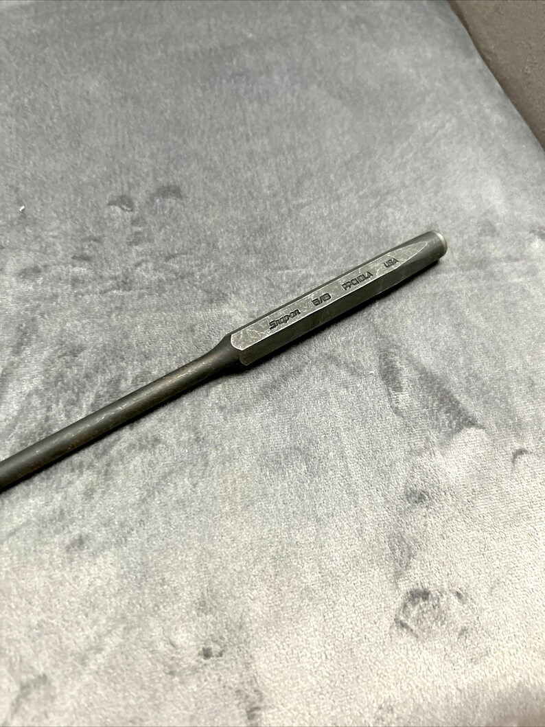 May include: A silver metal punch tool with the text "Snap-on 3/16" printed on the side. The tool is on a gray surface.