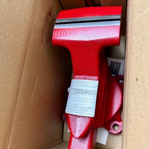 May include: A red metal vise with a silver jaw plate. The vise is in a cardboard box.
