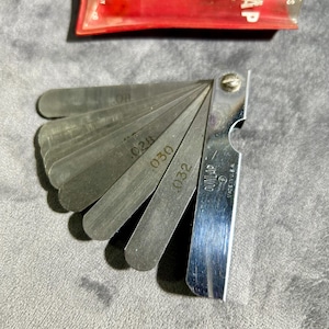 May include: A silver metal feeler gauge set with a red plastic case. The gauge has a fan of blades with different thicknesses marked in inches. The case has the text "Dunlap" and "Thickness Gauge" printed on it.