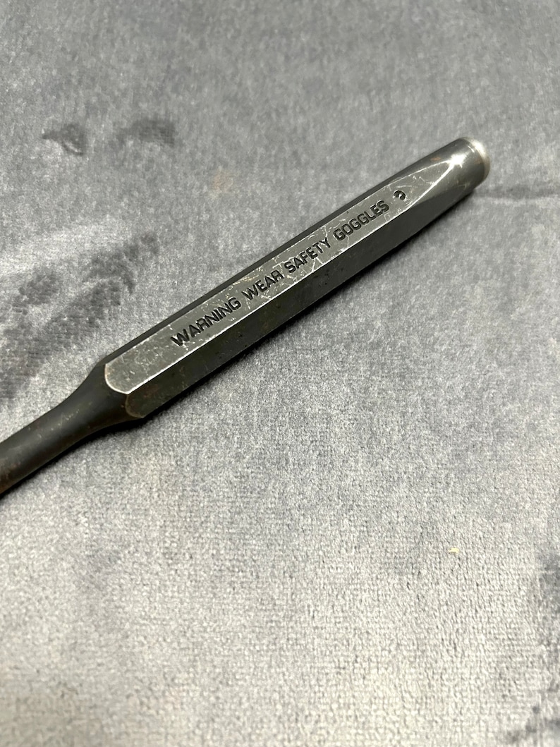 May include: A close-up of a metal punch tool with the text "WARNING WEAR SAFETY GOGGLES" engraved on the side.