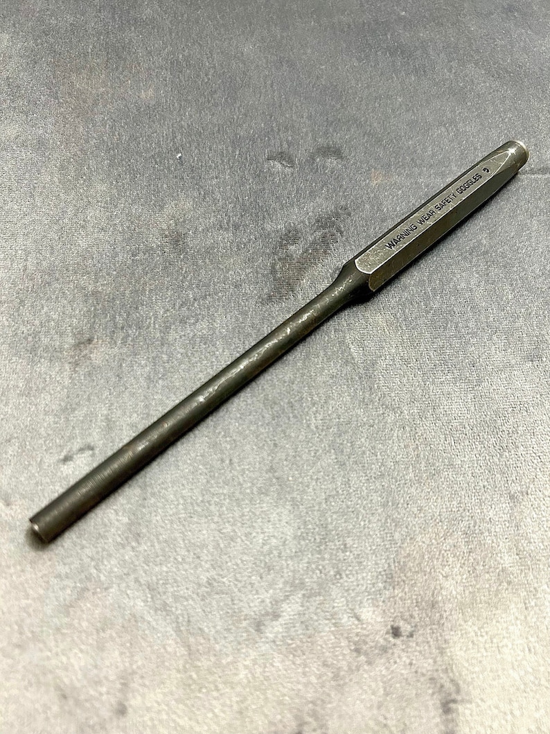 May include: A silver metal punch tool with a black handle. The tool has the text "WARNING WEAR SAFETY GOGGLES" printed on the side.