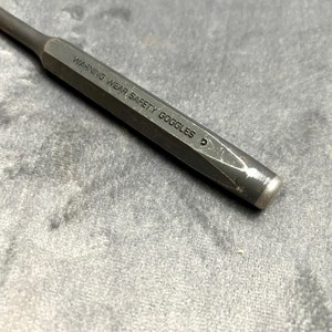 May include: A black metal punch with a hexagonal shaft and a pointed tip. The punch has the text "WARNING WEAR SAFETY GOGGLES" engraved on the side.