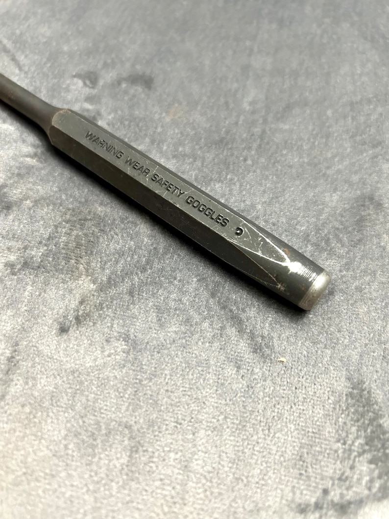 May include: A black metal punch with a hexagonal shaft and a pointed tip. The punch has the text "WARNING WEAR SAFETY GOGGLES" engraved on the side.