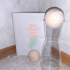Könnte beinhalten: Ein weißes Buch mit dem Titel "Die Kraft Der Zirbe" von Maximilian Moser, eine klare Glasflasche mit einem Holzstopfen und eine Holzkugel.