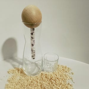 Könnte beinhalten: Eine klare Glaskaraffe mit einem hölzernen Kugelverschluss und einem kleinen Glas. Der Verschluss hat einen silbernen Ring und einen Stiel, der mit kleinen, dunklen Steinen gefüllt ist. Die Objekte befinden sich auf einer Oberfläche, die mit Holzspänen bedeckt ist.