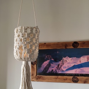 Peut inclure: Une lampe suspendue en macramé crème, avec un abat-jour cylindrique et une pampille. La lampe est suspendue à un cordon blanc et présente un motif tissé avec des espaces ouverts. En arrière-plan, un paysage de montagne encadré.
