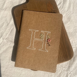 May include: A tan paper notebook featuring a large, white, hand-stitched letter H on the front cover. The letter is decorated with small, embroidered green leaves and a single pink flower bud stitched directly into the paper surface.