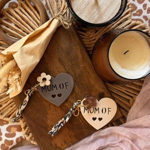 Peut inclure: Deux porte-clés en forme de cœur avec le texte "MUM OF" et deux découpes en forme de cœur. Un porte-clés est gris, l'autre est blanc cassé. Les deux ont un accent floral et une dragonne à motifs. Les porte-clés reposent sur une planche de bois.