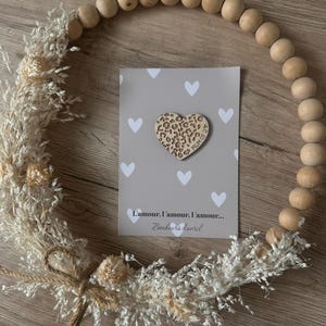 Peut inclure: Une carte de couleur gris clair avec une découpe en bois en forme de cœur blanche avec un motif léopard. La découpe porte l'inscription "Mum of 2". La carte porte l'inscription "L'amour. L'amour. L'amour... Bonheurs d'avril".