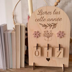 Peut inclure: Compteur de livres en bois avec le texte "Livres lus cette année". Le compteur comporte trois accents de fleurs roses et une illustration florale. Les chiffres "017" sont affichés sur de petites étiquettes en bois. Une pile de livres est visible à gauche.