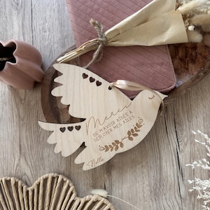 Peut inclure: Un ornement en bois en forme de colombe avec le texte français "Merci de m'avoir aidée à déployer mes ailes" et le nom "Nella". La colombe a des découpes en forme de cœur et est suspendue avec un ruban. Un dessous de verre matelassé rose et un bouquet de fleurs séchées sont également visibles.