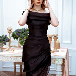 Puede incluir: Vestido negro con hombros descubiertos y falda drapeada. El vestido se lleva con joyas doradas. La imagen incluye el texto "KASHY KATY" en la parte inferior. El vestido es adecuado para eventos formales.