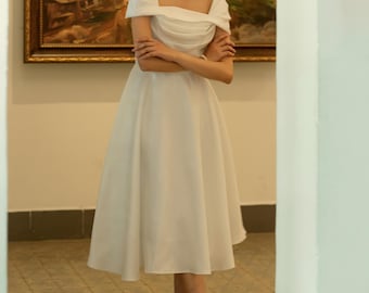 Puede incluir: Un vestido blanco hasta la rodilla con escote cuadrado y mangas descubiertas. El vestido se combina con tacones blancos y un sombrero a juego con velo. El texto "KASHY KATY" es visible en la parte inferior de la imagen.