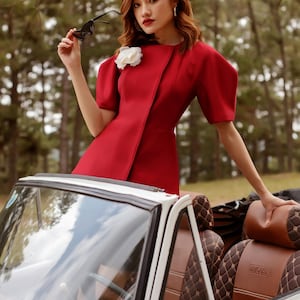 Può includere: Una donna con un vestito rosso e basco nero posa accanto a un'auto cabriolet bianca. L'abito ha maniche corte e un fiore bianco. L'interno dell'auto ha sedili in pelle marrone. La donna tiene occhiali da sole. Il testo "KASHY KATY" è visibile.