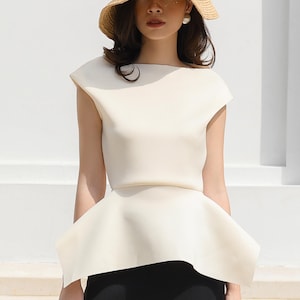 May include: A white peplum top with a boat neck and short sleeves. The top is paired with a black skirt and a straw hat with a black ribbon that says "JADIOR".