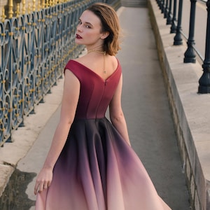 Op de afbeelding: Een formele jurk met een kleurengradatie, van bordeauxrood naar zwart en vervolgens naar licht perzik. De off-the-shoulder halslijn en de volle rok cre&euml;ren een elegante silhouet. Een gouden ketting voegt een vleugje glamour toe. De merknaam "KASHY KATY" is zichtbaar.