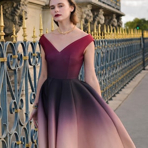Op de afbeelding: Een mouwloze, knielange jurk met een V-hals en een ombre-effect, dat overgaat van bordeauxrood naar zwart en vervolgens naar lichtroze. De jurk wordt gedragen met een gouden ketting en armband. De tekst "KASHY KATY" is onderaan zichtbaar.