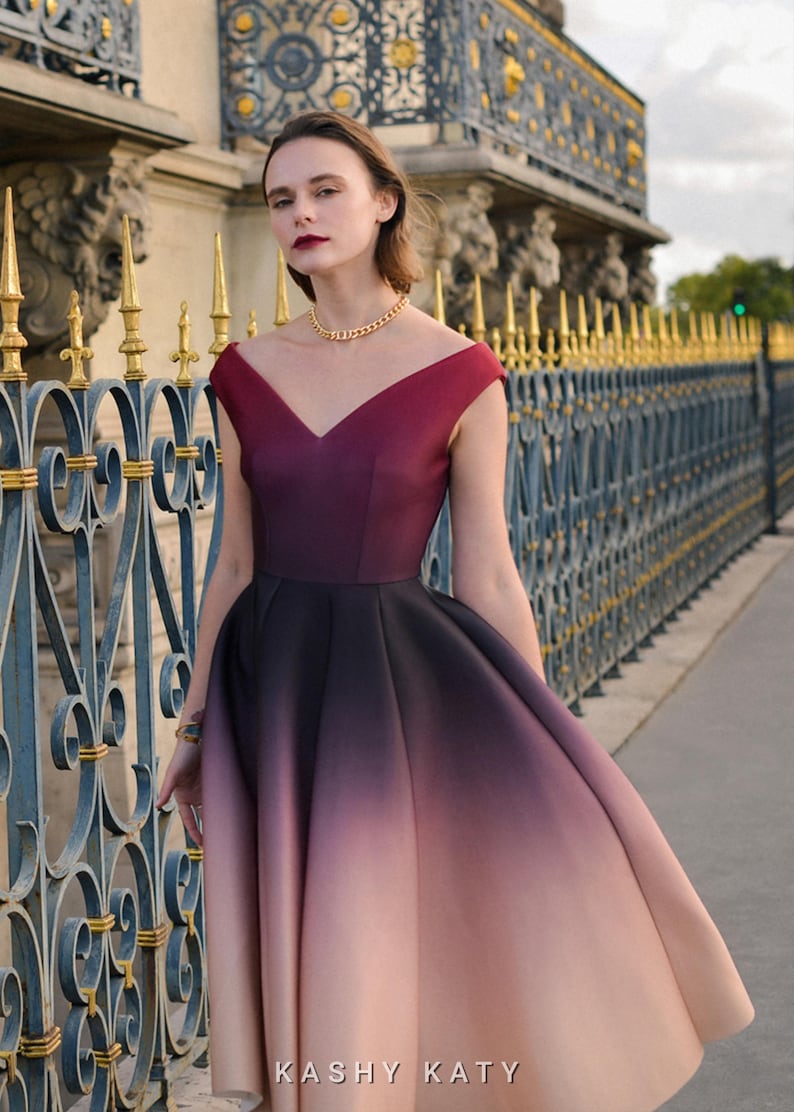 Op de afbeelding: Een mouwloze, knielange jurk met een V-hals en een ombre-effect, dat overgaat van bordeauxrood naar zwart en vervolgens naar lichtroze. De jurk wordt gedragen met een gouden ketting en armband. De tekst "KASHY KATY" is onderaan zichtbaar.