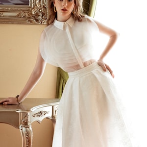 Könnte beinhalten: Weißes zweiteiliges Outfit mit einem transparenten Oberteil mit Kragen und einem knielangen Rock. Das Oberteil hat ein geschichtetes Design und der Rock einen zarten Spitzenbesatz. Der Markenname KASHY KATY ist sichtbar.
